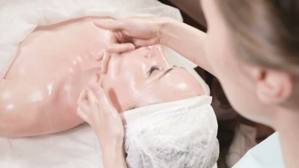Chica esteticista en una máscara y guantes hace un masaje facial saludable a una mujer atractiva. Masaje cosmetológico de nueva generación — Vídeos de Stock