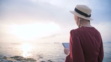 Arkadan bakıldığında yaz aylarında kırmızı elbiseli ve hasır şapkalı çekici bir kız gün batımında deniz kenarında bir taşın üzerinde oturup tablette bir şeyler izliyor. Ekran boyunca kaydır