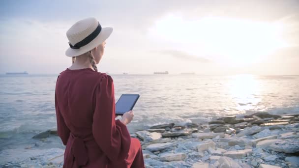 Vista pelas costas Uma jovem atraente em um vestido vermelho de verão e um chapéu de palha se senta em uma pedra junto ao mar ao pôr do sol e observa algo em um tablet. Passe o dedo pela tela — Vídeo de Stock