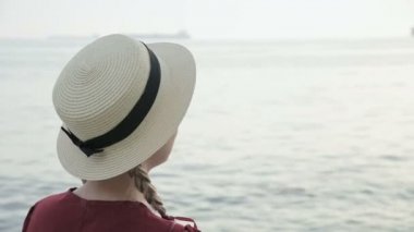 Yazın kırmızı elbisesi ve hasır şapkası olan beyaz bir kızın güneş batarken sahile oturmuş şapkasını düzeltiyor.