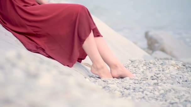 Kırmızı elbiseli ve at kuyruklu hasır şapkalı çekici beyaz kadın deniz kıyısında oturur kameraya bakarak gülümser. — Stok video