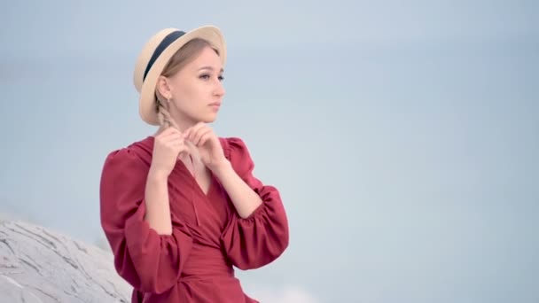 Ritratto attraente giovane donna caucasica in abito rosso e un cappello di paglia con le trecce si gira seduto sulla riva del mare sorridente guardando la fotocamera contro il mare in sfocatura — Video Stock