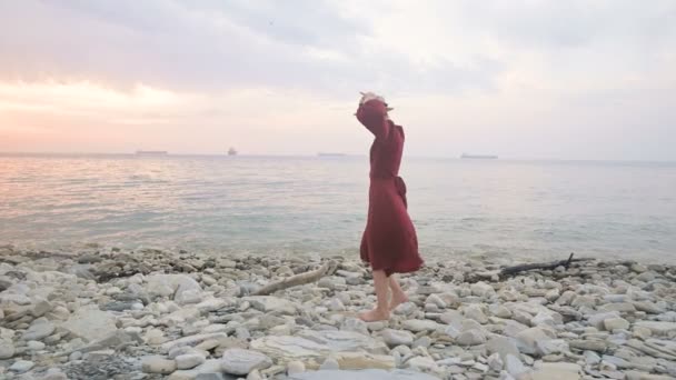 Una joven atractiva con un vestido rojo de verano y un sombrero de paja se arrastra cautelosamente sobre las piedras de la costa rocosa del mar contra el fondo de las olas y la puesta de sol . — Vídeos de Stock