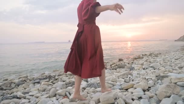 En attraktiv ung flicka i en röd sommarklänning och en halmhatt försiktigt kryper på stenarna i havet stenig strand mot bakgrund av vågor och solnedgång. — Stockvideo