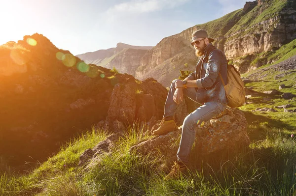Közelkép egy szakállas férfiról farmerben, napszemüvegben, sapkában, hátizsákkal az epikus sziklák lábánál. Divat jellegű — Stock Fotó