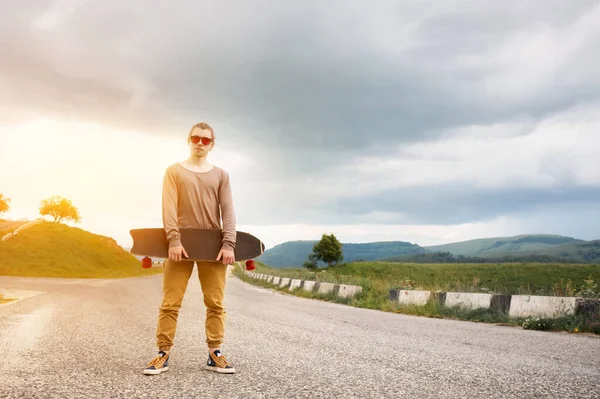 Egy stílusos fiatalember áll egy kanyargós hegyi úton, egy rája vagy Longboard a kezében este naplemente után. Az ifjúsági sport-és utazási hobbi — Stock Fotó
