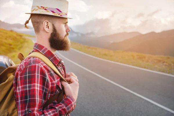 Retrato de un barbudo feliz viajero serio hipster con una mochila en una camisa a cuadros y un sombrero al lado de un coche desconocido se encuentra en la carretera al atardecer en las montañas. Concepto de viaje feliz y seguro — Foto de Stock