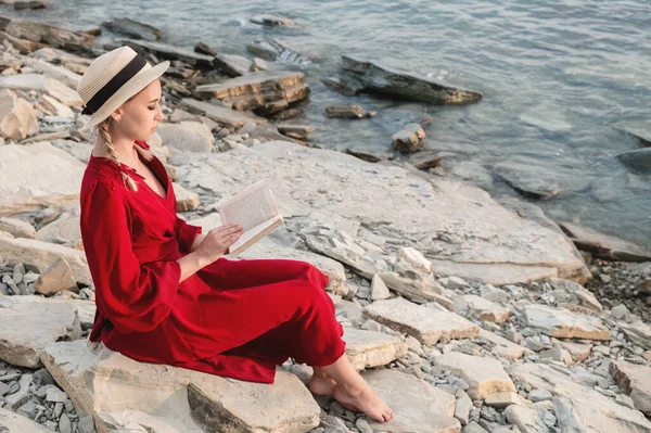 Atraktivní Kavkazská dívka v červených, světlých šatech a slaměném klobouku sedí na velkém kameni u moře při západu slunce a čte zajímavou knihu o mořském obzoru a nádherné mraky. — Stock fotografie