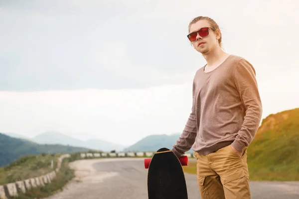 Um jovem elegante de pé ao longo de uma estrada de montanha sinuosa com um skate ou longboard em suas mãos na noite após o pôr do sol. O conceito de esportes juvenis e hobbies de viagem — Fotografia de Stock