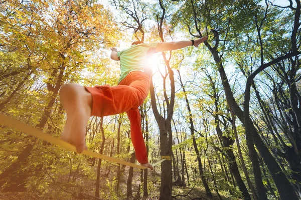 Wide angle male tightrope walker balancing barefoot on slackline in autumn forest. The concept of outdoor sports and active life of people aged