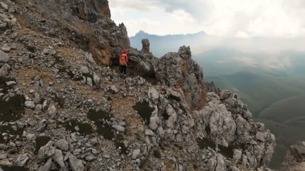 Aerial view of a girl traveler with a backpack stands on the edge of a cliff of a high rocky mountain and looks at the snow-capped mountains in the distance. Travel video drone footage — Stock Video