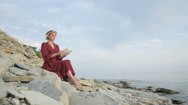 Atrakcyjna kaukaski dziewczyna w czerwonej jasnej sukni i kapelusz słomy siedzi na dużym kamieniu nad morzem o zachodzie słońca i czyta ciekawą książkę na horyzoncie morskim i pięknych chmur. — Wideo stockowe