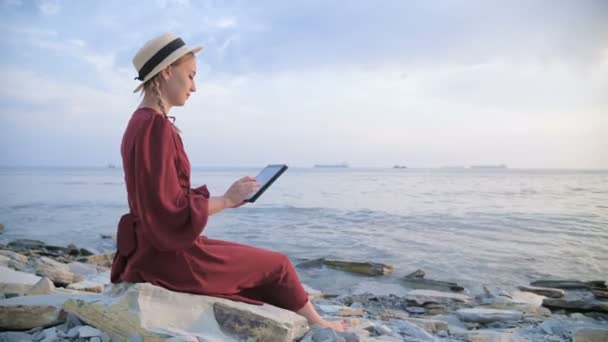 Uma jovem atraente em um vestido vermelho de verão e um chapéu de palha se senta em uma pedra à beira-mar à noite e olha para algo em um tablet. Passe o dedo pela tela — Vídeo de Stock
