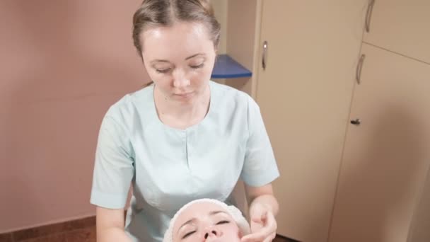Chica esteticista en una máscara y guantes hace un masaje facial saludable a una mujer atractiva. Masaje cosmetológico de nueva generación — Vídeo de stock