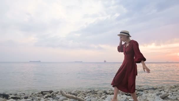 Una joven atractiva con un vestido rojo de verano y un sombrero de paja se arrastra cautelosamente sobre las piedras de la costa rocosa del mar contra el fondo de las olas y la puesta de sol . — Vídeos de Stock