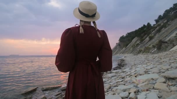 La vista desde atrás. Retrato en cámara lenta de una joven con un vestido rojo y un sombrero de paja en la orilla del mar. Una chica en el viento sostiene un sombrero con sus manos. Esperando el regreso de los marineros — Vídeos de Stock