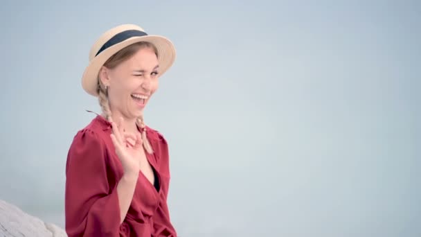 Portrét atraktivní bělošky v červených šatech a slamáku s copánky sedícími na břehu, usmívající se na kameru a ukazující ok gesto proti moři v rozostření — Stock video