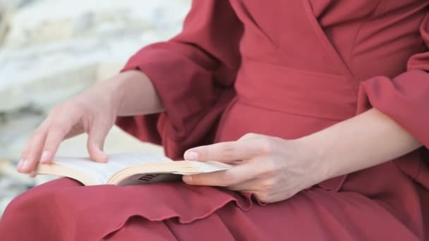 Close-up uma menina caucasiana atraente mãos em um vestido vermelho brilhante e um chapéu de palha senta-se em uma grande pedra junto ao mar ao pôr do sol e lê um livro interessante — Vídeo de Stock