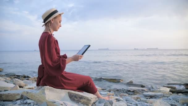 O tânără atractivă într-o rochie roșie de vară și o pălărie de paie stă pe o piatră lângă mare seara și se uită la ceva pe o tabletă. Trageți cu degetul pe ecran — Videoclip de stoc