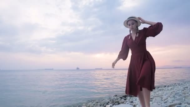 Egy vonzó fiatal lány piros nyári ruhában és szalmakalapban óvatosan kúszik a köveken a tenger sziklás partján a hullámok és naplemente hátterében.. — Stock videók