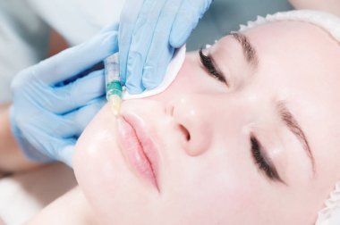 Yakın plan çekici genç kadın yaşlanma karşıtı yüz enjeksiyonları yaptırıyor. Bir klinikte ya da kuaförde sakince yatıyor. Deneyimli genç bir kozmetikçi kadın kırışıklıklarını şırınganın hyaluronik asidiyle dolduruyor.