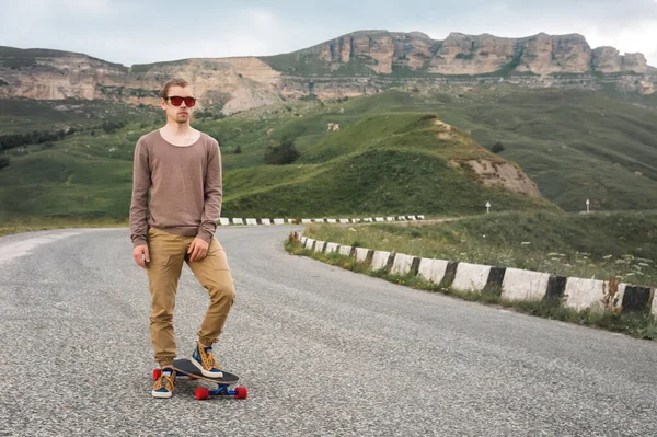 Ein stylischer junger Mann, der am Abend nach Sonnenuntergang mit Skate oder Longboard in der Hand an einer kurvenreichen Bergstraße steht. das Konzept des Jugendsports und der Reiselust — Stockfoto
