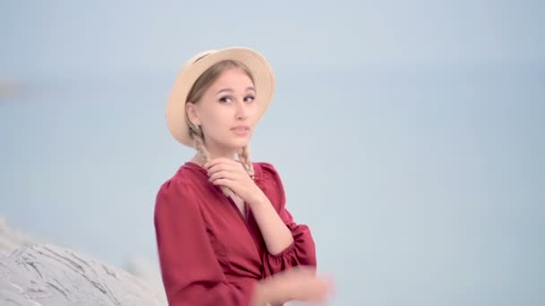 Портрет приваблива біла молода жінка в червоній сукні і солом'яний капелюх з кісками обертається, сидячи на березі моря, посміхаючись дивитися на камеру проти моря в дефокусі — стокове відео