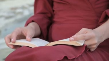 Yakın çekim kırmızı parlak elbise ve hasır şapka lı çekici beyaz bir kız gün batımında denizin yanında büyük bir taşın üzerinde oturur ve ilginç bir kitap okur