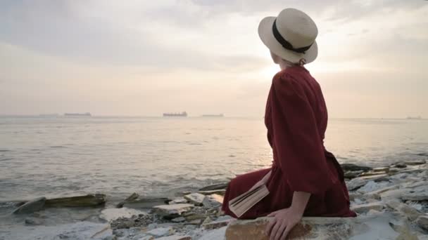 Приваблива кавказька дівчина в червоній яскравій сукні і солом'яний капелюх сидить на великому камені біля моря на заході і читає цікаву книгу про морську горизонт і прекрасні хмари. — стокове відео