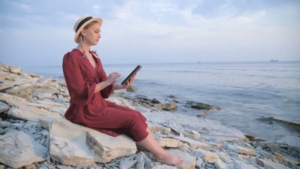 Привлекательная молодая девушка в летнем красном платье и соломенной шляпе вечером сидит на камне у моря и смотрит на что-то на планшете. Прокатиться по экрану — стоковое видео