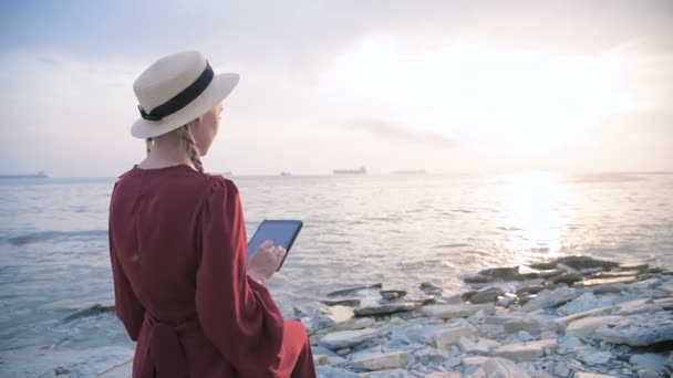 Arkadan bakıldığında yaz aylarında kırmızı elbiseli ve hasır şapkalı çekici bir kız gün batımında deniz kenarında bir taşın üzerinde oturup tablette bir şeyler izliyor. Ekran boyunca kaydır — Stok video