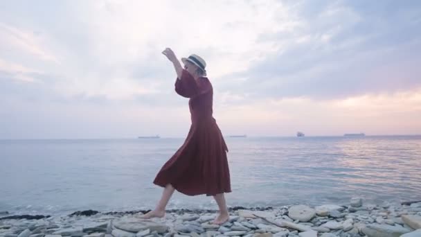 Ένα ελκυστικό νεαρό κορίτσι με ένα κόκκινο καλοκαιρινό φόρεμα και ένα ψάθινο καπέλο σέρνεται προσεκτικά στις πέτρες της βραχώδους ακτής της θάλασσας στο φόντο των κυμάτων και του ηλιοβασιλέματος. — Αρχείο Βίντεο