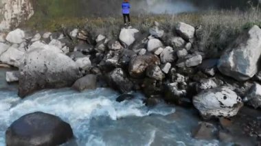 Bir kız nehir kenarındaki bir taşın üzerinde duran ve büyük destansı bir şelaleyi fotoğraflayan profesyonel bir manzara fotoğrafçısı. Hava görüntüsü. Kuzey Kafkasya