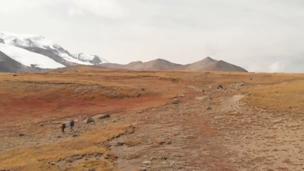 Büyük sırt çantalı, şapkalı ve güneş gözlüklü bir çift gezginin havadan görünüşü destansı dağlarla çevrili bir plato boyunca yürüyor. — Stok video