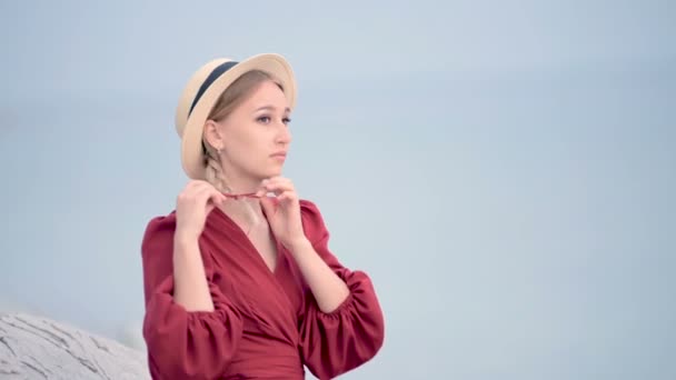 Portrait attractive caucasian young woman in a red dress and a straw hat with pigtails turns around sitting on the seashore smiling looking at the camera against the sea in defocus — Stock Video