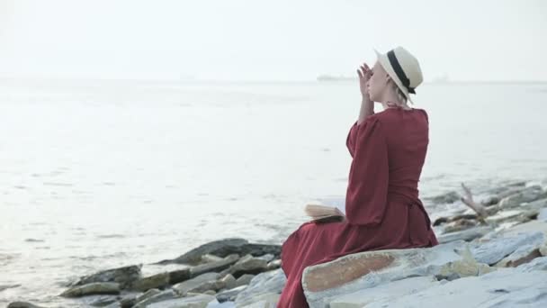 Indietro Una bella ragazza caucasica in un abito rosso brillante e un cappello di paglia siede su una grande pietra in riva al mare al tramonto e legge un libro interessante sull'orizzonte del mare e belle nuvole . — Video Stock