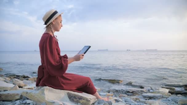 An attractive young girl in a summer red dress and a straw hat sits on a stone by the sea in the evening and looks at something on a tablet. Swipe across the screen — Stock Video