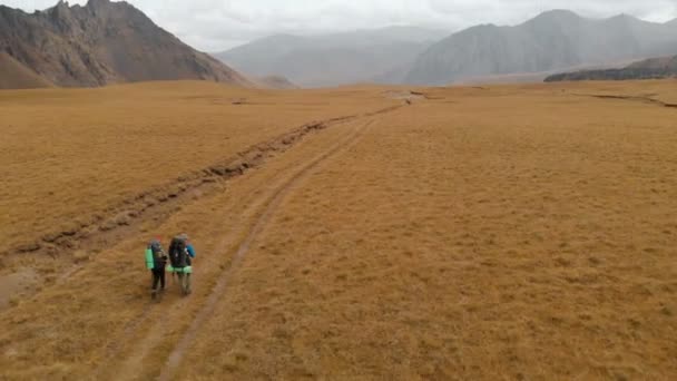 Aerial view of a couple of travelers man and woman with large backpacks in hats and sunglasses walk along an alpine plateau surrounded by epic mountains — Stock Video