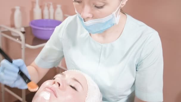 Flickan en kosmetolog utför proceduren att rengöra och återfukta huden. Applicera en mask med en speciell borste för ett kvinnligt ansikte. — Stockvideo