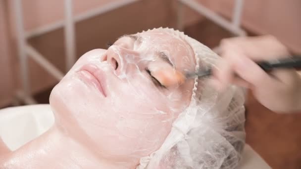 A lány kozmetológus elvégzi az eljárást a bőr tisztítására és hidratálására. Vigyen fel egy maszkot egy speciális kefével egy női arcra. Bőrápolás. — Stock videók