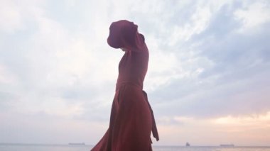 Kırmızı bir yaz elbisesi ve hasır şapkası olan çekici genç bir kız denizin taşlarında dikkatle sürünüyor dalgaların ve günbatımının arka planında kayalık kıyılarda.