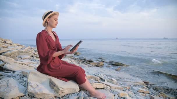 Una joven atractiva con un vestido rojo de verano y un sombrero de paja se sienta en una piedra junto al mar por la noche y mira algo en una tableta. Desliza el dedo por la pantalla — Vídeos de Stock