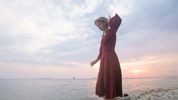 Una joven atractiva con un vestido rojo de verano y un sombrero de paja se arrastra cautelosamente sobre las piedras de la costa rocosa del mar contra el fondo de las olas y la puesta de sol . — Vídeo de stock