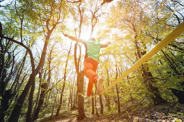 Wide angle male tightrope walker balancing barefoot on slackline in autumn forest. The concept of outdoor sports and active life of people aged