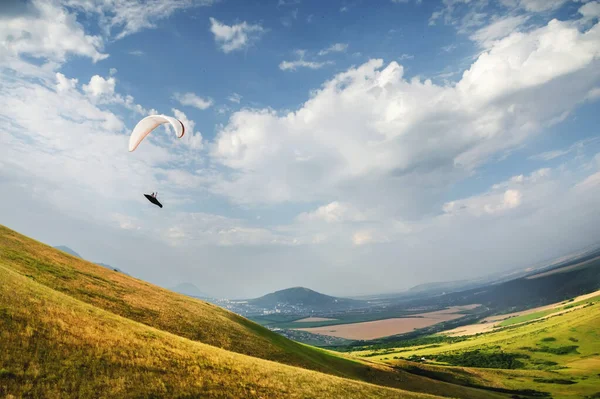 Un parapente vuela en el cielo en un traje de capullo en un parapente sobre el campo caucásico con colinas y montañas. Parapente Concepto Deportivo — Foto de Stock