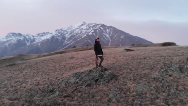 Arial pohled stylově oblečené lyžařské oblečení dívka v čepici stojí na vysokém kopci večer po západu slunce modrá hodina a má výhled na hory. — Stock video