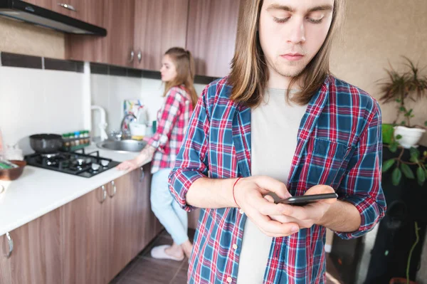 Serious long-haired young man standing in the kitchen uses his mobile phone to write SMS or order a service from the Internet — Stock Photo, Image