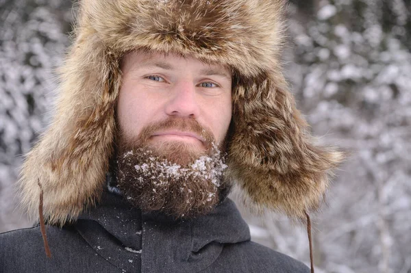 Retrato de um homem barbudo em um chapéu grande pele contra o fundo de uma floresta coberta de neve. Homem viril sorridente na floresta — Fotografia de Stock