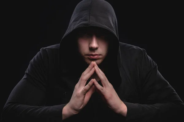 A low-key studio portrait of a large man in a hood sits pondering. Sex faces in the dark hacker stealth and anonymous surveillance hidden concept