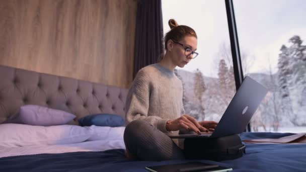 Uma mulher freelancer com óculos senta-se em uma cama em uma casa de campo com janelas panorâmicas atrás das quais há uma floresta nevada. Menina com computador portátil faz remotamente trabalho enquanto isolado — Vídeo de Stock